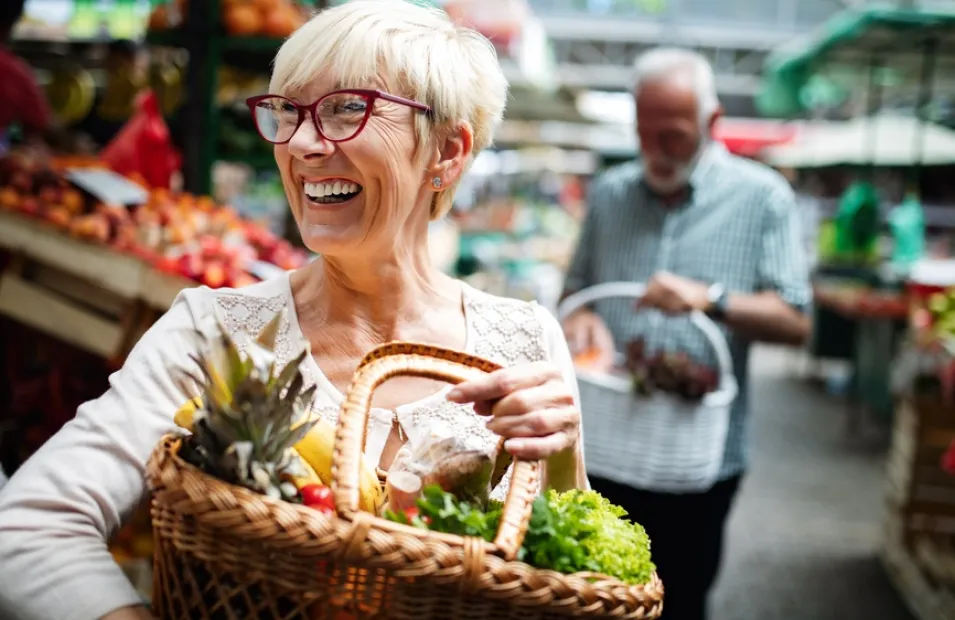 Older smiling woman holding a basket of fruit and vegetables at a market