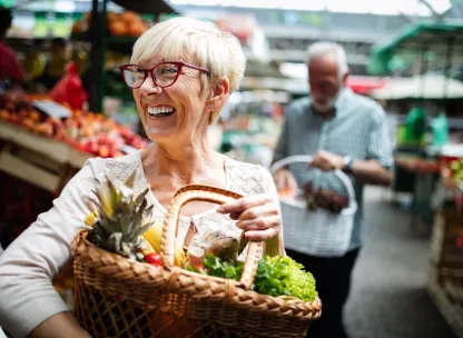 Older smiling woman holding a basket of fruit and vegetables at a market