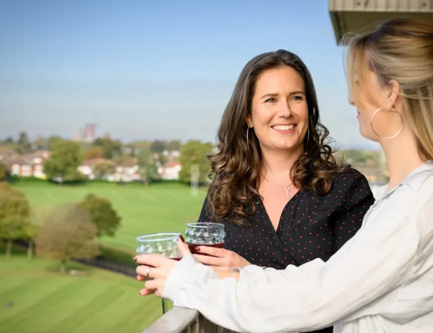 Two smiling women standing on a balcony drinking wine overlooking green fields